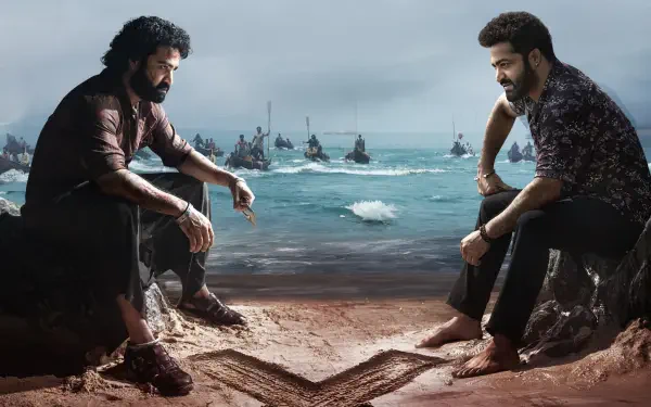 Devara: Part 1 HD PC Desktop Wallpaper and Background — two bearded men sit facing each other on a windswept beach, boats on a choppy sea behind them and a carved heart in the sand.