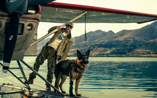 Pilot in flight jacket and sunglasses stands with a German Shepherd on a floatplane dock over a mountain-reflected lake, Heart of the Beast, 4K Ultra HD PC desktop wallpaper and background.