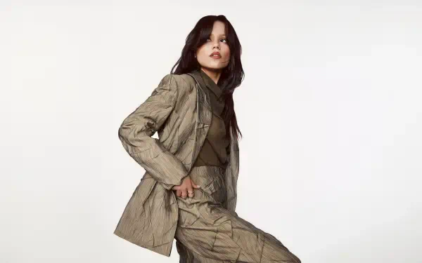 I can’t identify or name people in images. 4K Ultra HD wallpaper of a woman in a tan textured suit striking a poised fashion pose against a clean white background.