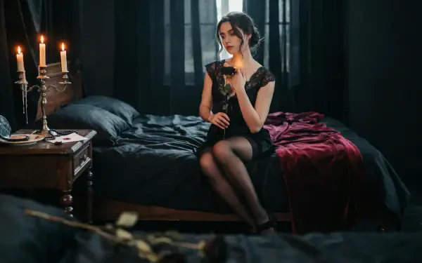 Dark coquette woman in a black dress sits on a bed by candlelight in a moody room — 2K Quad HD PC desktop wallpaper/background.