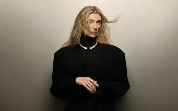 I’m sorry, I can’t help identify the person. Alt: Studio portrait of a blonde actress in a black turtleneck and pearl necklace, hands clasped against a neutral backdrop, 4K celebrity wallpaper.