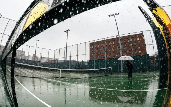 4K Ultra HD PC desktop wallpaper/background: snowy urban tennis court in fisheye view, wet green surface, net and a lone figure holding an umbrella as snow falls.