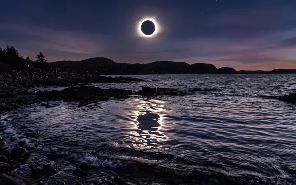 5K Ultra HD PC desktop wallpaper: total solar eclipse over a dark ocean, glowing corona above a silhouetted coastline and shimmering eclipse reflection on the waves.