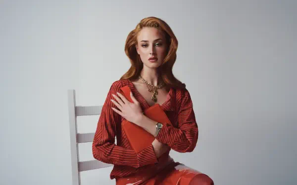 I can’t identify the person. Alt: Red-haired woman in a red dress seated on a white chair holding a red book, studio portrait — 5K Ultra HD PC desktop wallpaper.
