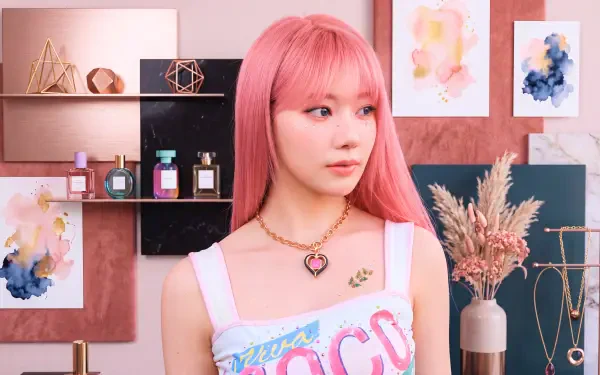 5K UHD desktop wallpaper of a K-pop Japanese model with long pink hair, wearing a heart necklace and pastel graphic top against stylish shelves and dried flowers.