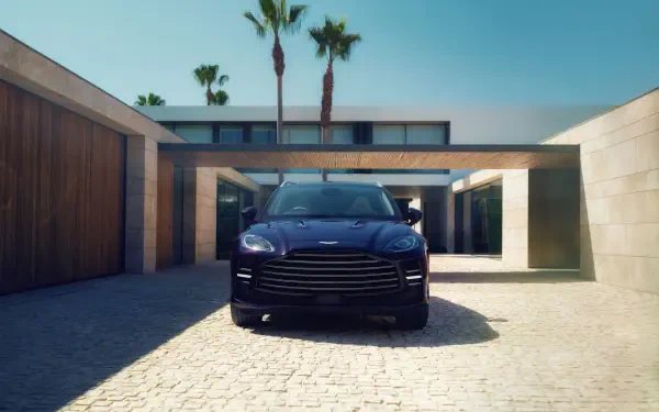 Front view of a blue Aston Martin DBX GT SUV in a sunlit modern courtyard — a luxury car presented as a 2K Quad HD PC desktop wallpaper.