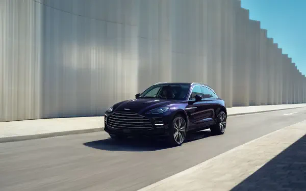 Aston Martin DBX GT sedan driving past modern concrete walls, captured as a 2K Quad HD PC desktop wallpaper and background.