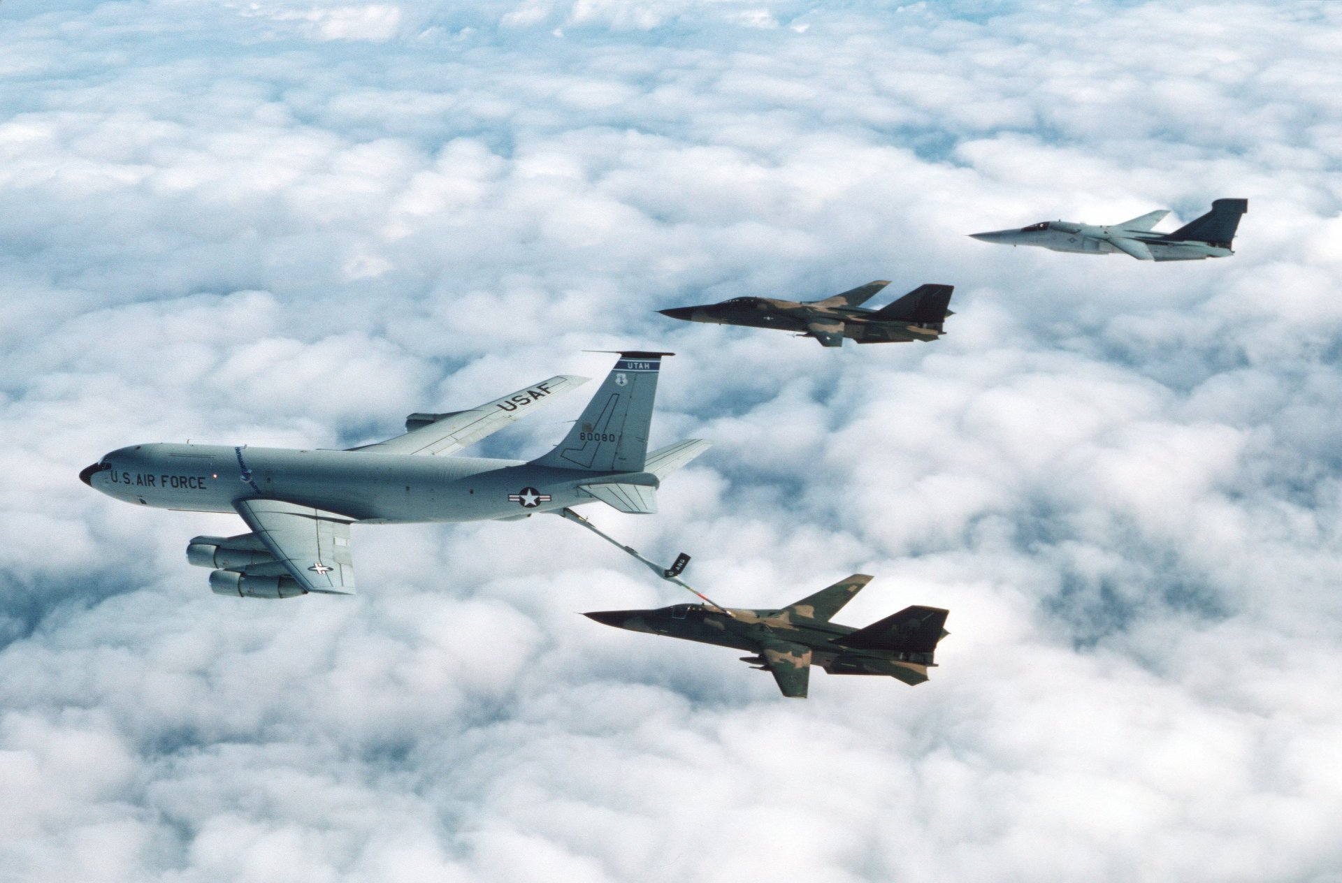Aerial view of a Boeing KC-135 Stratotanker refueling two General Dynamics F-111 Aardvark jet fighters flying above a cloud layer, captured in HD desktop wallpaper quality.