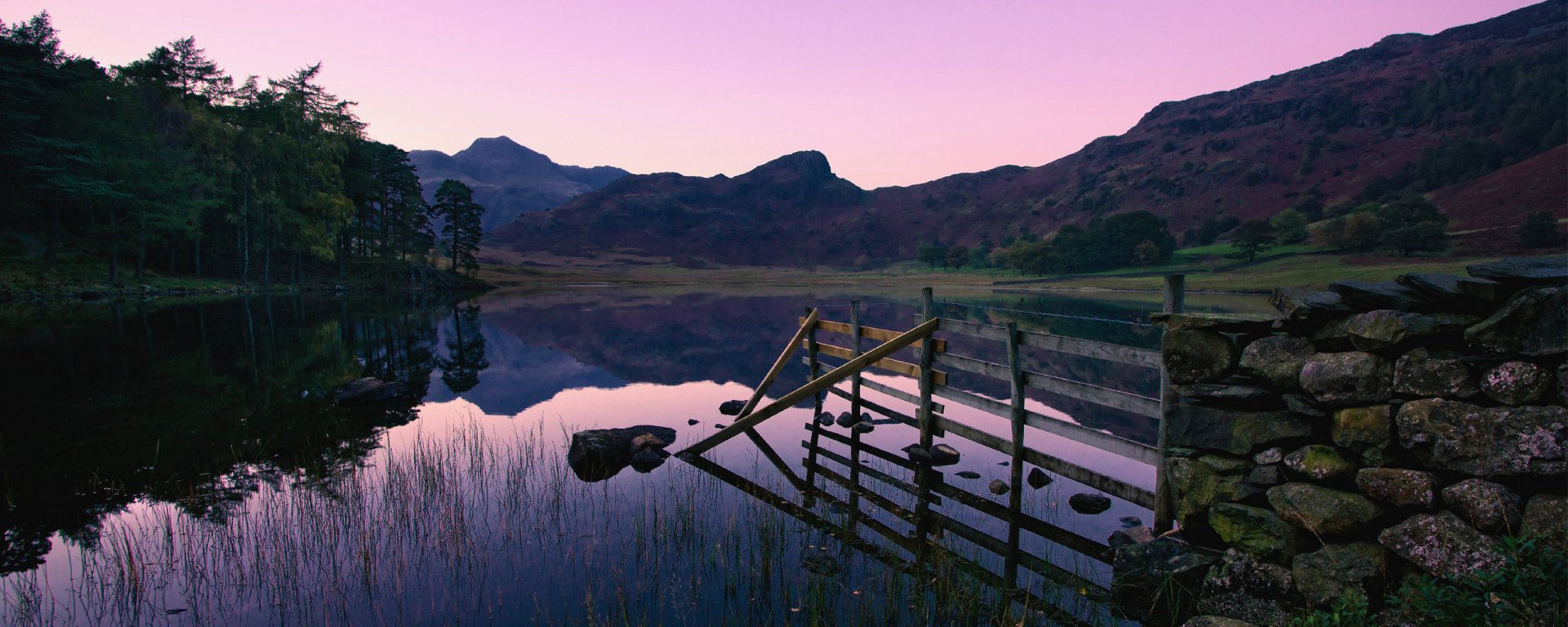 HD photography of a serene lake at dusk, featuring calm water reflecting mountains and a wooden fence, framed by trees, captured as a PC desktop wallpaper background.