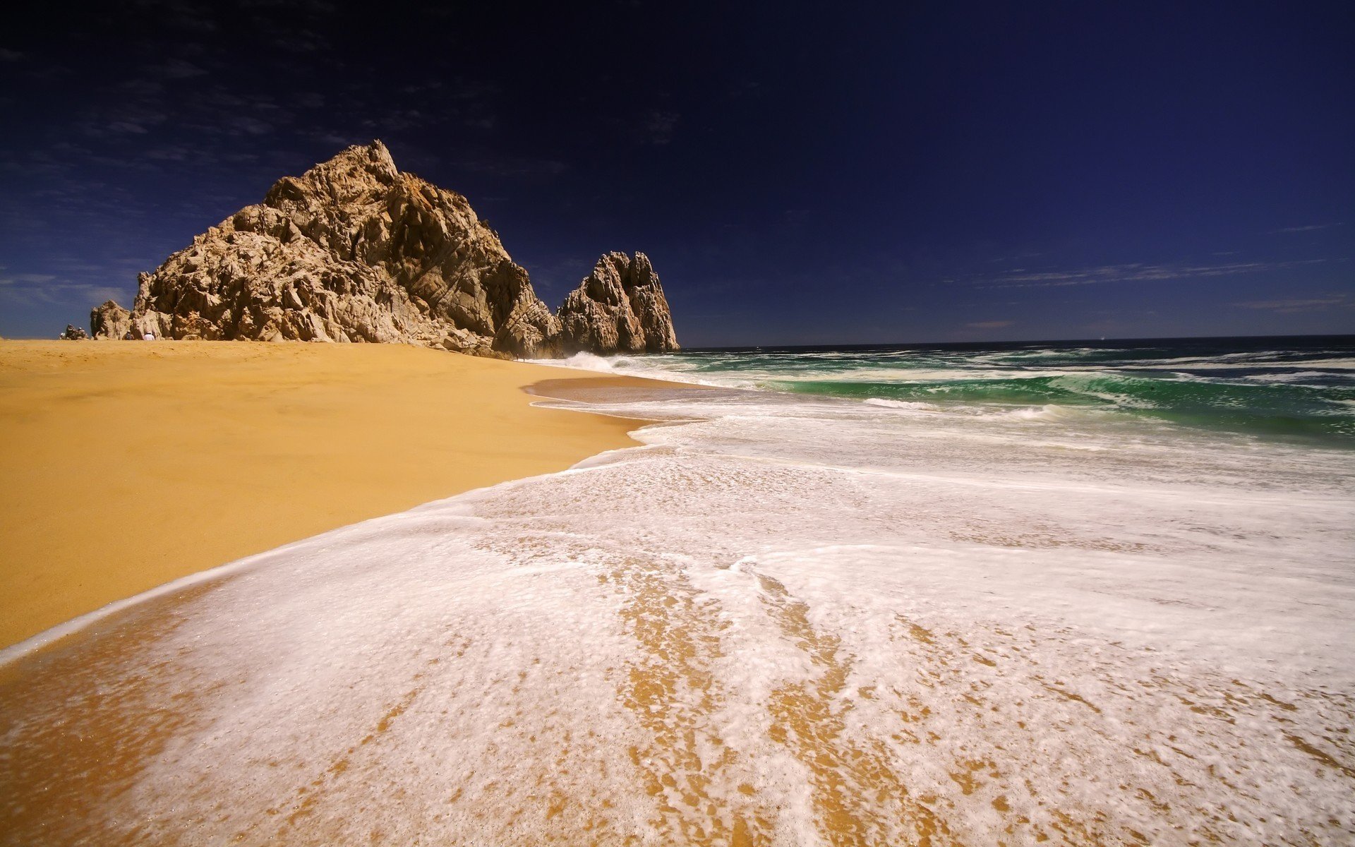 HD desktop wallpaper capturing ocean waves gently washing over sandy beach with rocky formations under a deep blue sky, showcasing nature’s serene beauty.