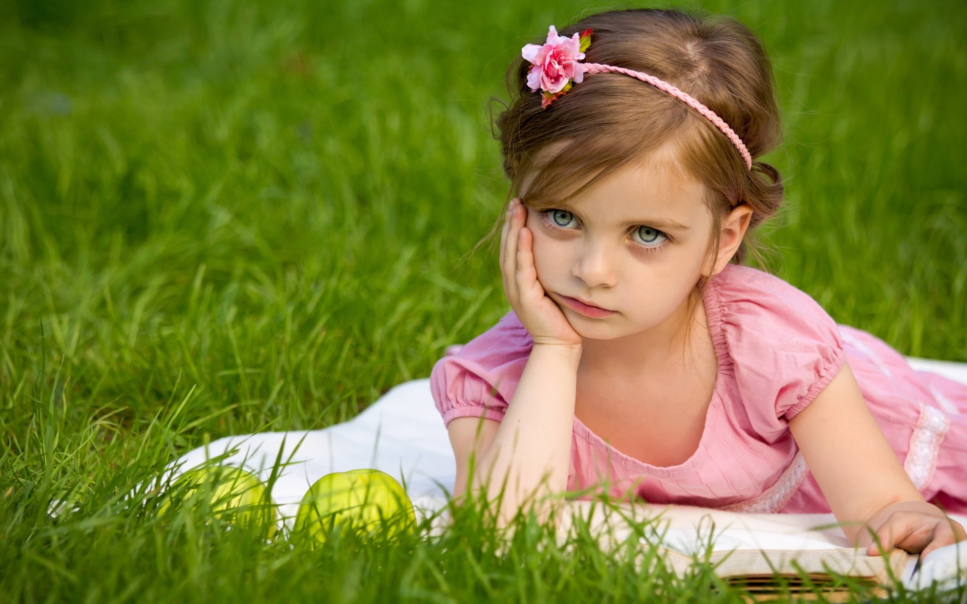 A young girl with a floral headband lies on a white blanket in green grass, resting her chin on her hand. Her thoughtful expression captures a serene moment in this HD photography image.