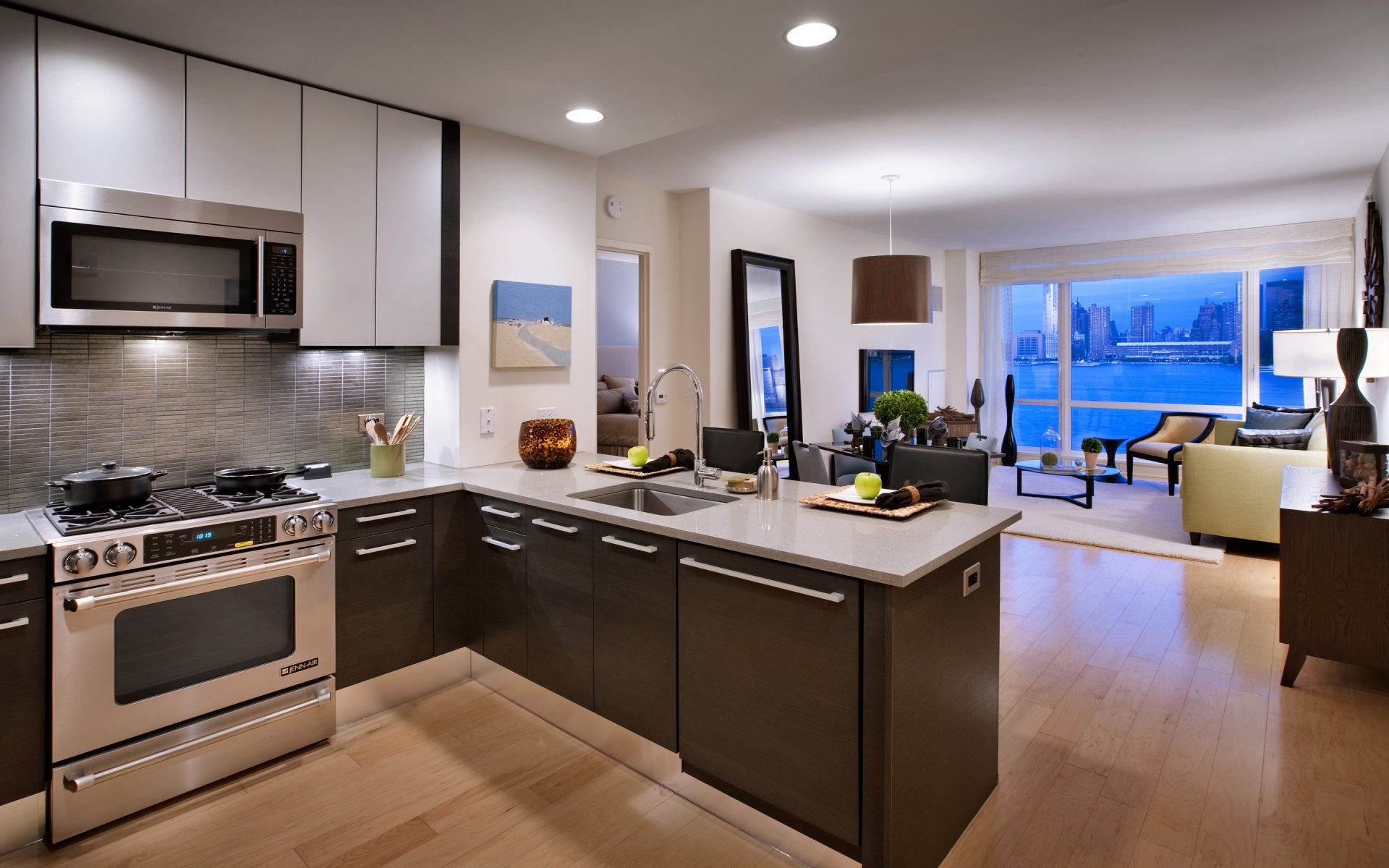 HD desktop wallpaper showing a modern man-made kitchen with sleek cabinetry, stainless steel appliances, and an open layout extending to a living room with large windows and city views.