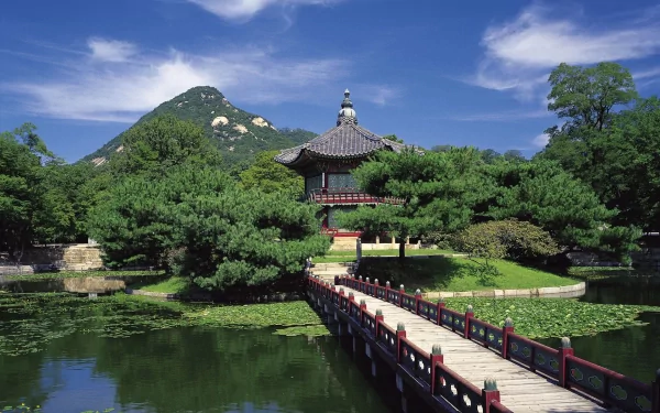 Scenic view of a traditional Korean shrine on Jeju Island, South Korea, with a wooden bridge over water and lush greenery under a clear blue sky.