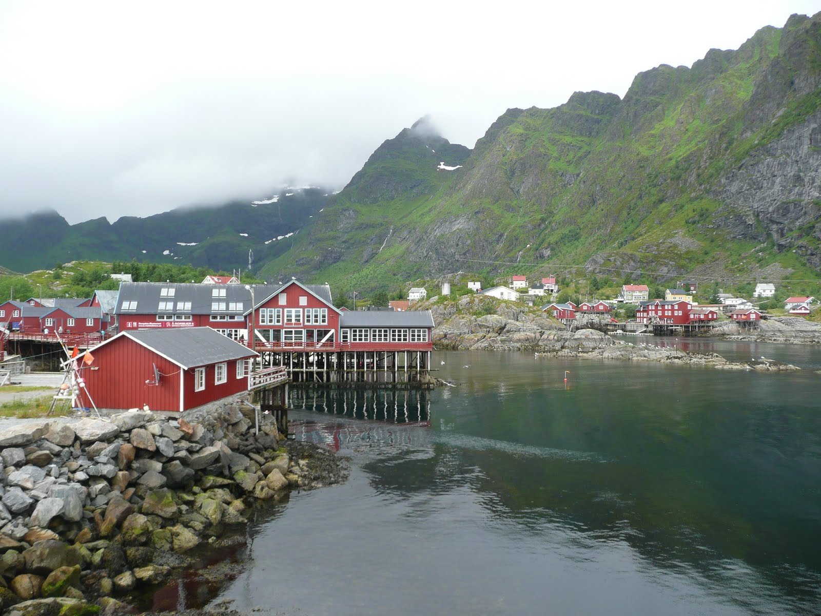 HD PC desktop wallpaper/background showing a man-made coastal town of red wooden houses on stilts along a rocky shore and calm fjord, green mountains rising in mist.
