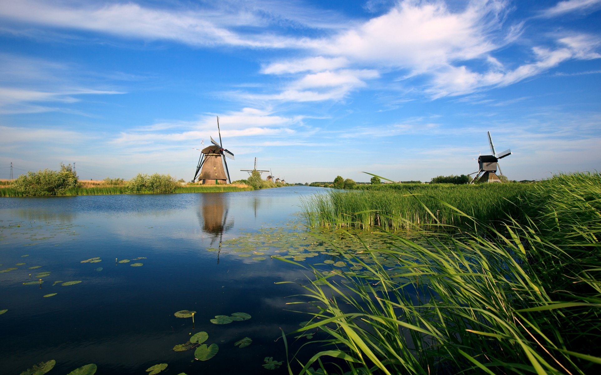 HD PC desktop wallpaper featuring serene countryside with man-made windmills reflected in a calm water body under a bright blue sky with scattered clouds.