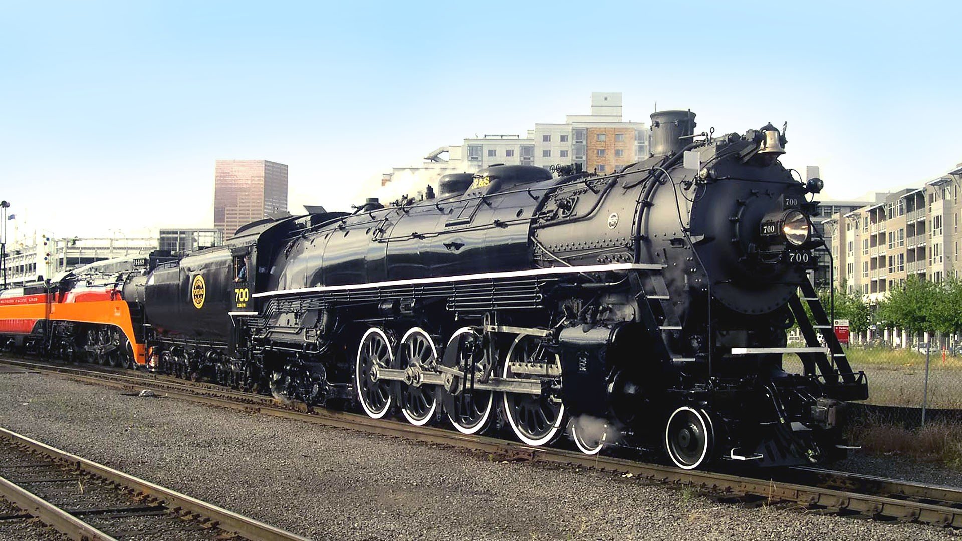 A powerful steam train stands on the tracks, showcasing its intricate detailing against a backdrop of modern buildings, making it a striking HD PC desktop wallpaper and background.