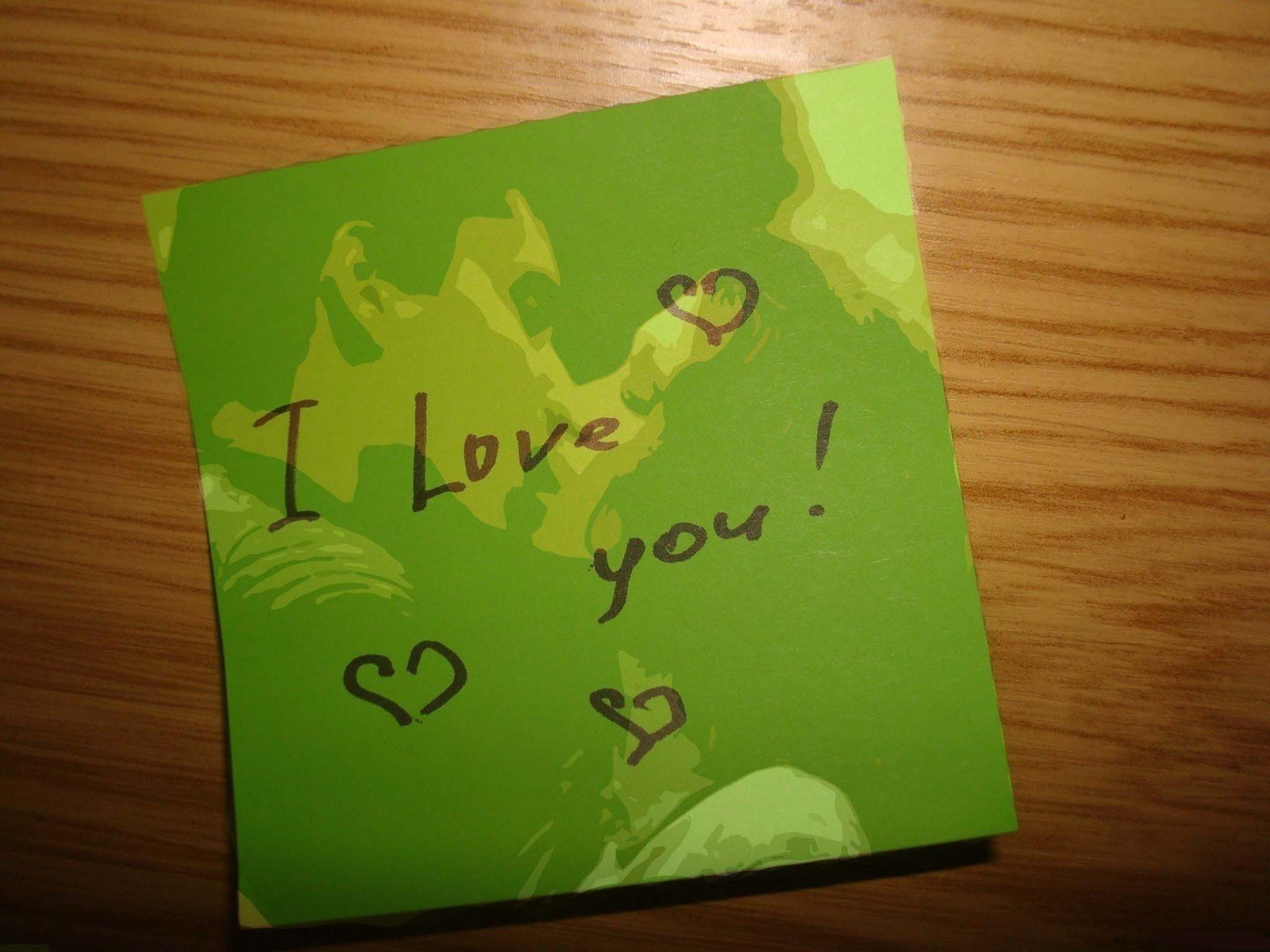 A vibrant green sticky note features the message I Love you! accompanied by heart doodles, set against a wooden background, creating an artistic representation of love.