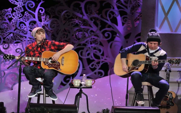 HD wallpaper featuring Justin Bieber and another musician sitting on stools, playing acoustic guitars in a festive, snowy setting. The background includes intricate, decorative snowflakes.