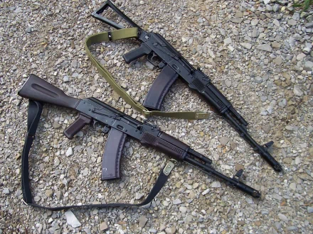 HD PC desktop wallpaper: two man-made AKM assault rifles with slings resting on gravel.