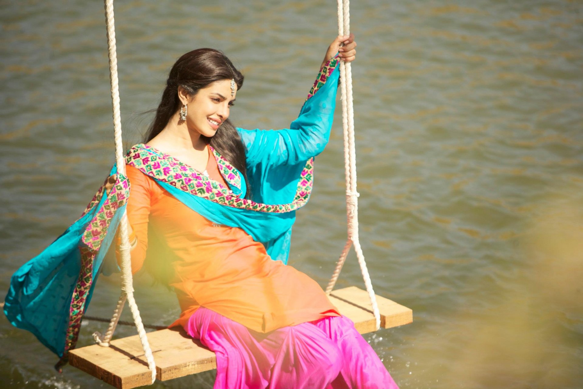 Woman in a vibrant traditional outfit on a wooden swing over calm water, smiling — HD PC desktop wallpaper/background.