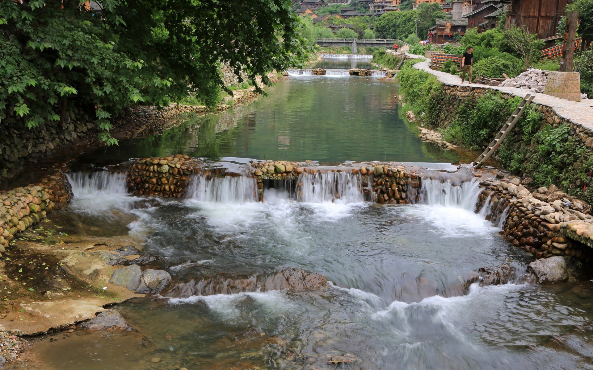 Photography of a tranquil riverside place with stone weirs and a footbridge, emerald water flowing past village houses — HD PC Desktop Wallpaper and Background.