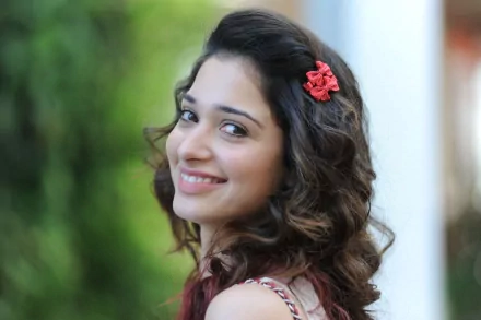 A vibrant HD wallpaper featuring the celebrity Tamannaah Bhatia, smiling and looking over her shoulder, complemented by a floral hair accessory and a blurred green background.