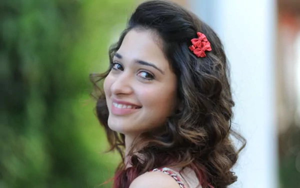 A vibrant HD wallpaper featuring the celebrity Tamannaah Bhatia, smiling and looking over her shoulder, complemented by a floral hair accessory and a blurred green background.