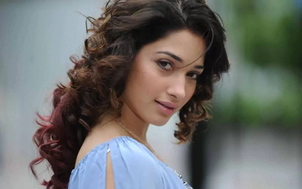 HD desktop wallpaper featuring celebrity Tamannaah Bhatia with soft curls and a light blue outfit against a blurred outdoor background.
