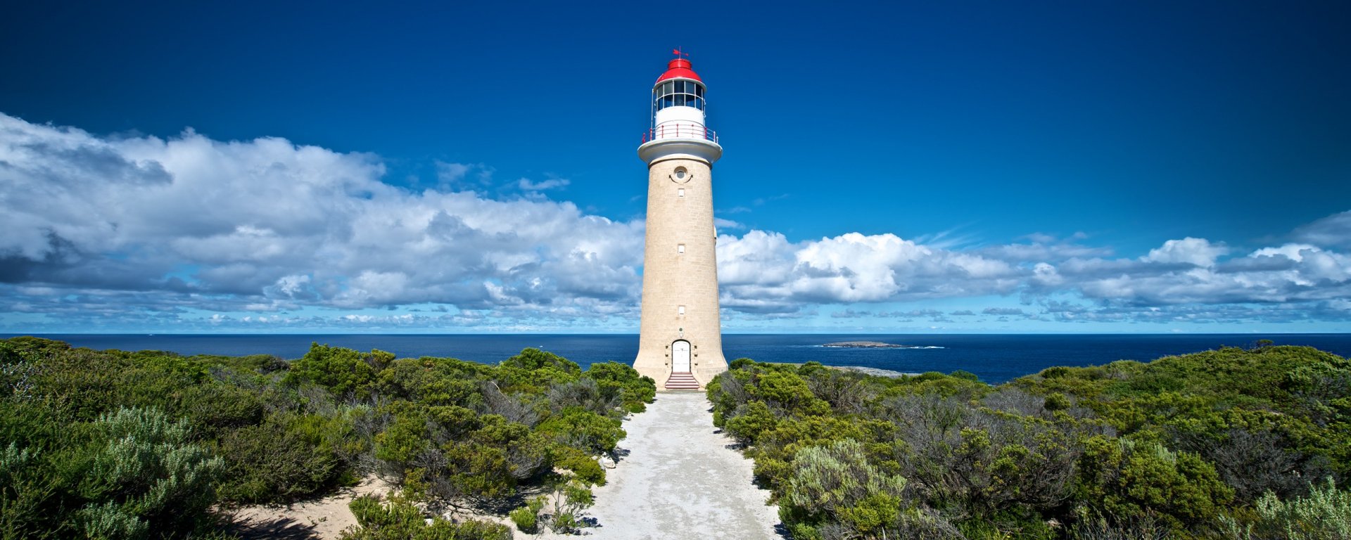 HD PC desktop wallpaper of a man-made Australian lighthouse on a coastal headland, bright white tower with red lantern, pathway through green scrub toward the blue ocean and sky.