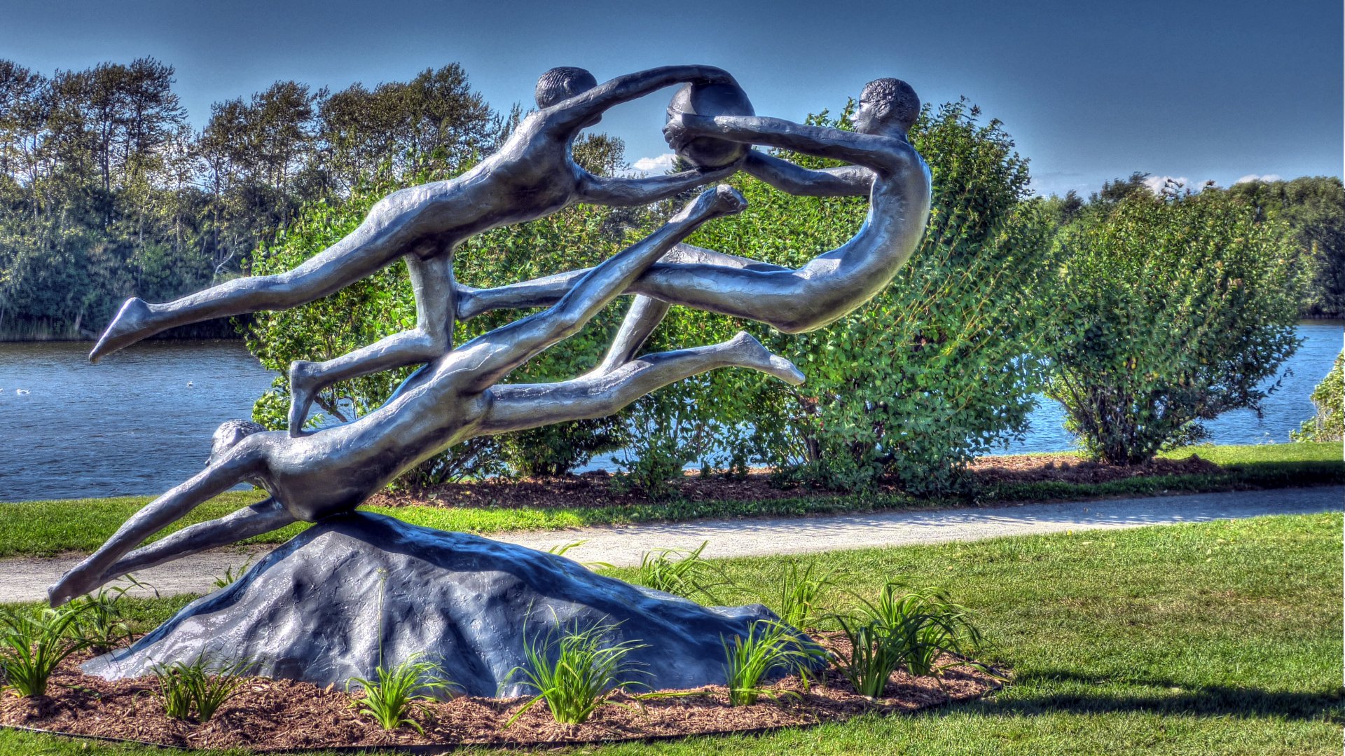 4K Ultra HD wallpaper featuring a dynamic man-made statue of three intertwined human figures in a park setting with lush greenery and a calm river background.
