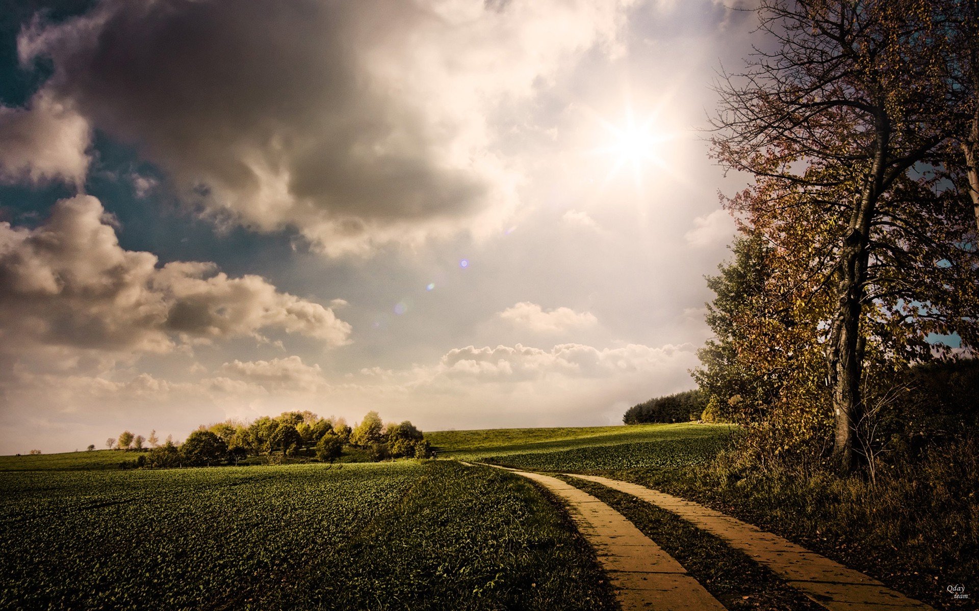 HD PC desktop wallpaper showing a sunlit green field with a winding dirt path, trees to the side and a dramatic cloud-filled sky with the bright sun breaking through.