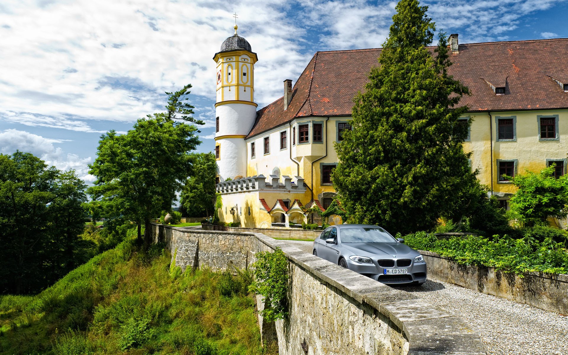 HD PC desktop wallpaper featuring a sleek BMW vehicle parked on a stone driveway in front of a historic castle surrounded by lush greenery under a partly cloudy sky.
