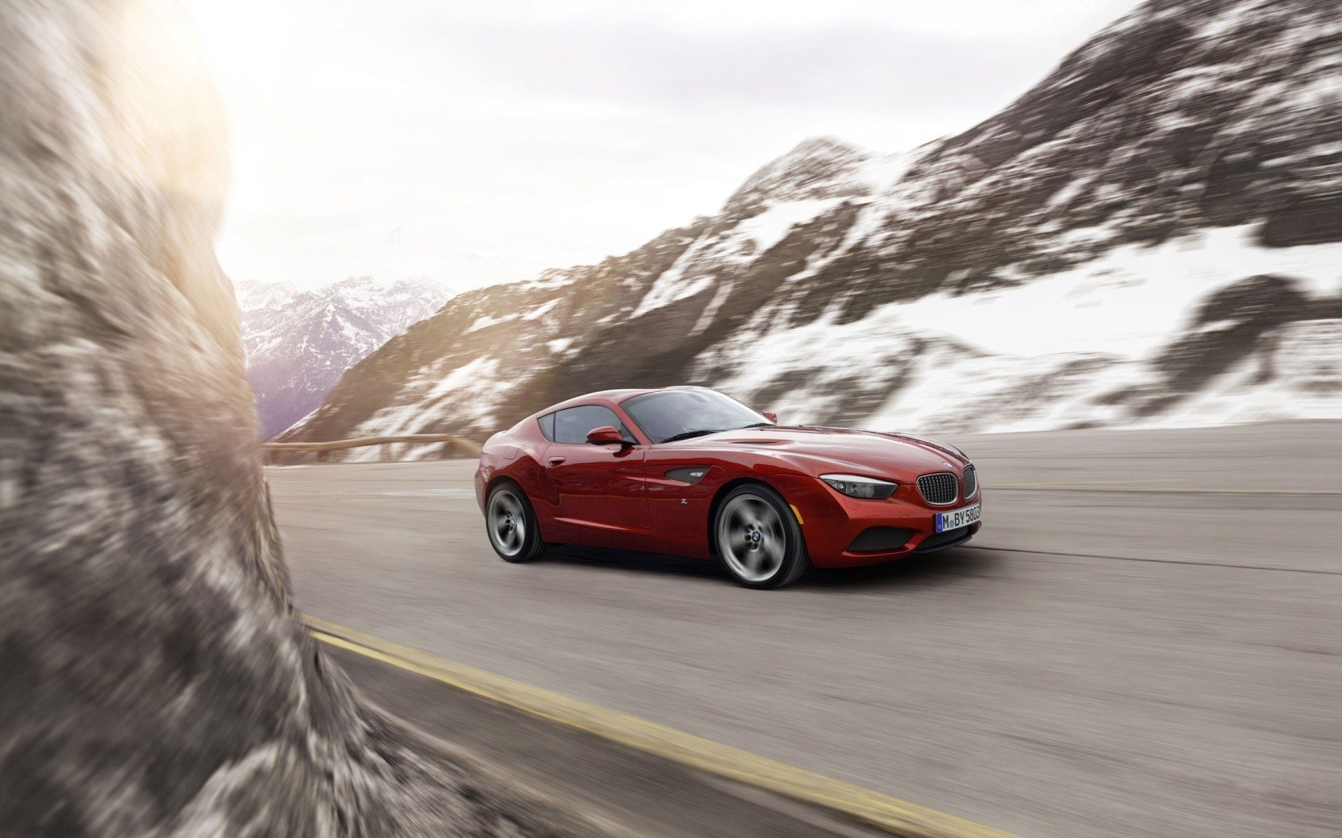 HD desktop wallpaper featuring a sleek red BMW sports car driving on a mountain road with snowy peaks in the background.