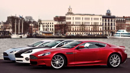 A stunning HD wallpaper showcasing a sleek red Aston Martin sports car alongside two other luxury vehicles, set against a picturesque waterfront cityscape.
