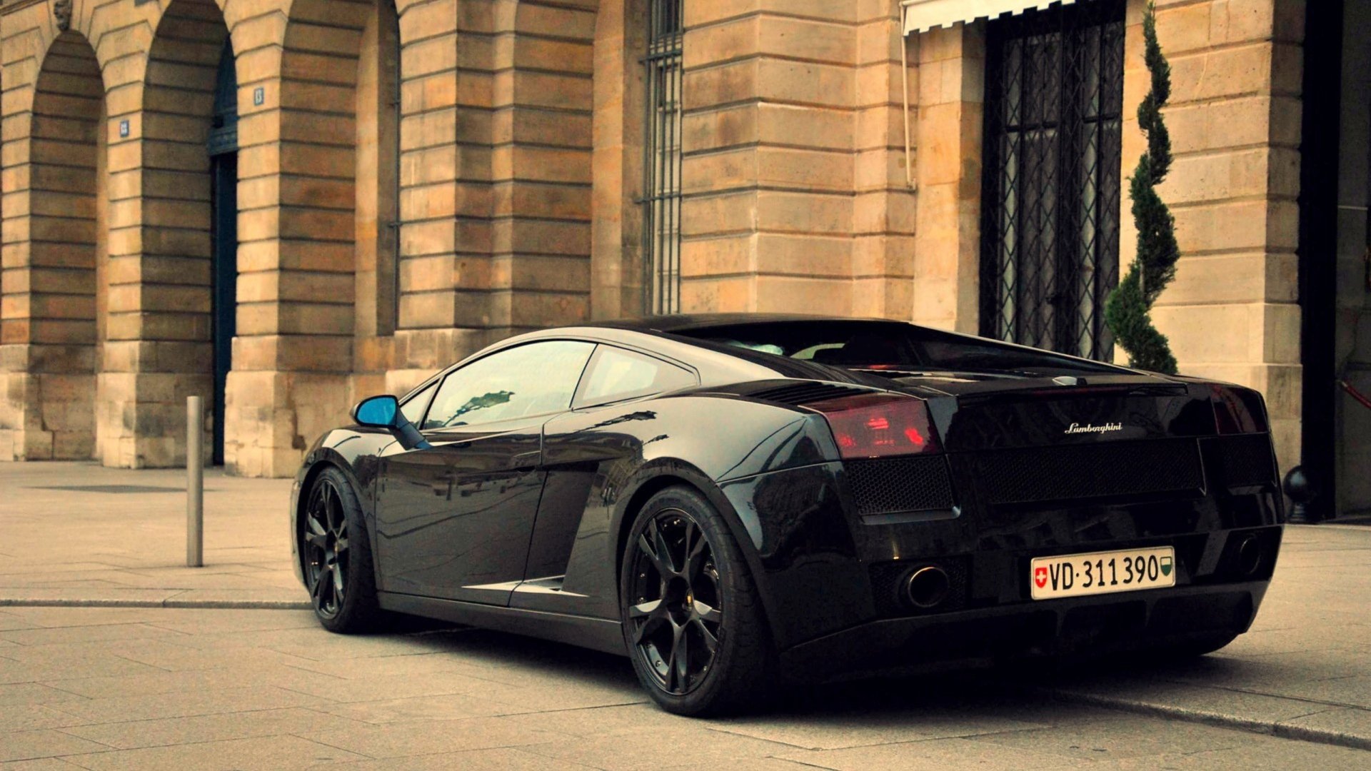 HD PC desktop wallpaper of a black Lamborghini Gallardo vehicle parked on a city street, sleek low profile and glossy finish against a stone-arched building backdrop.