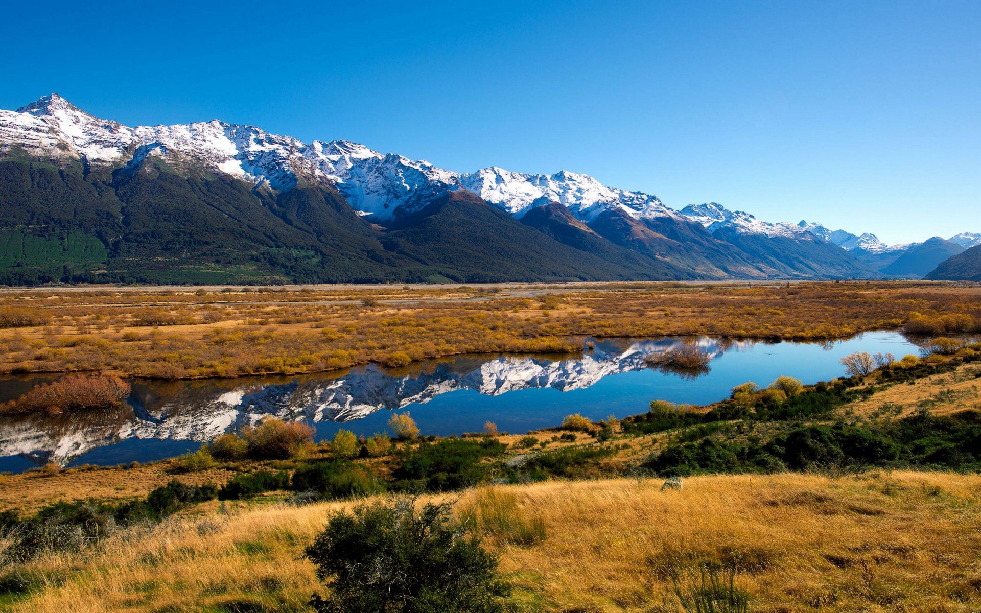 A scenic HD desktop wallpaper featuring a river reflecting snow-capped mountains, with a serene lake and natural landscape under a clear blue sky.