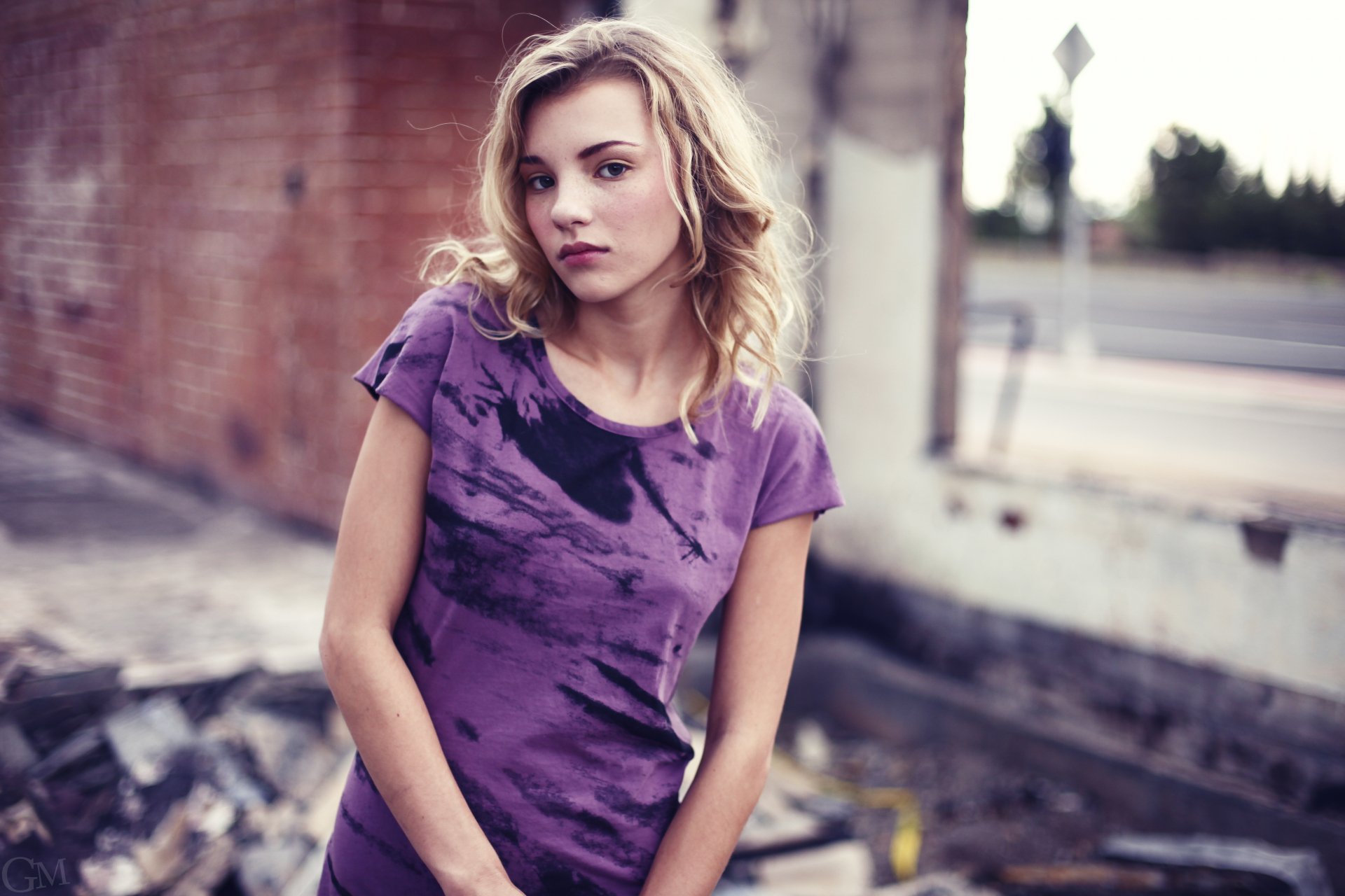 Portrait of a woman with blonde hair wearing a purple tie-dye shirt, set against an urban background, captured in 4K Ultra HD by Rachel Ann Yampolsky.