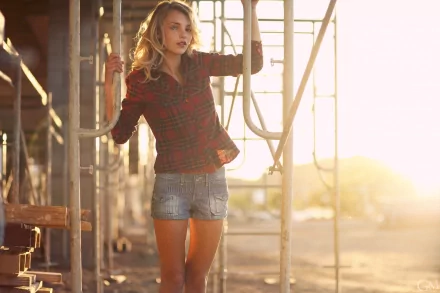 A blonde woman named Rachel Ann Yampolsky stands in sunlight wearing shorts and a plaid shirt, set against a scaffold backdrop. This HD wallpaper captures a serene outdoor moment.