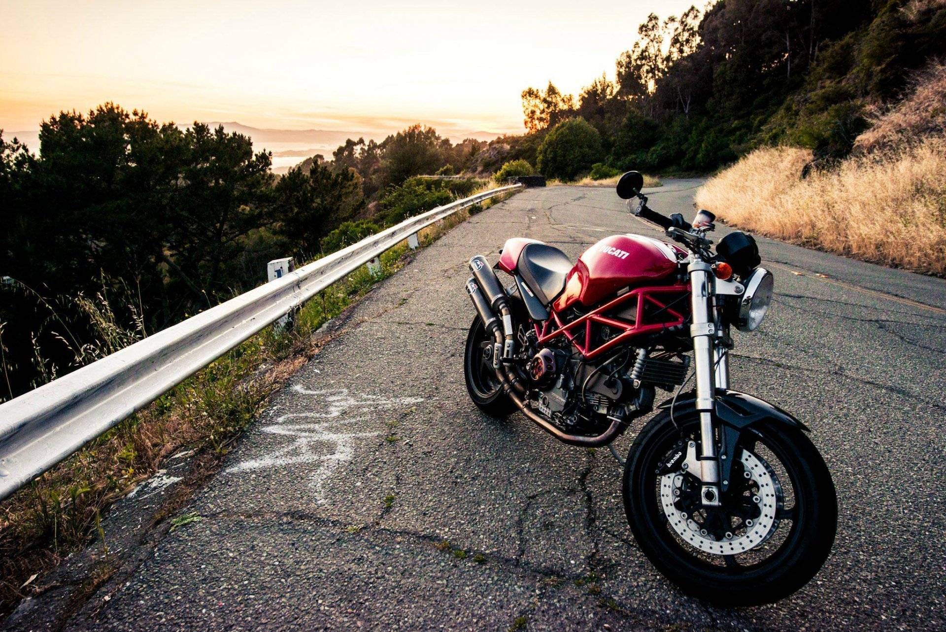 HD PC desktop wallpaper featuring a red Ducati motorcycle parked on a scenic winding road at sunset with trees and guardrails in the background.