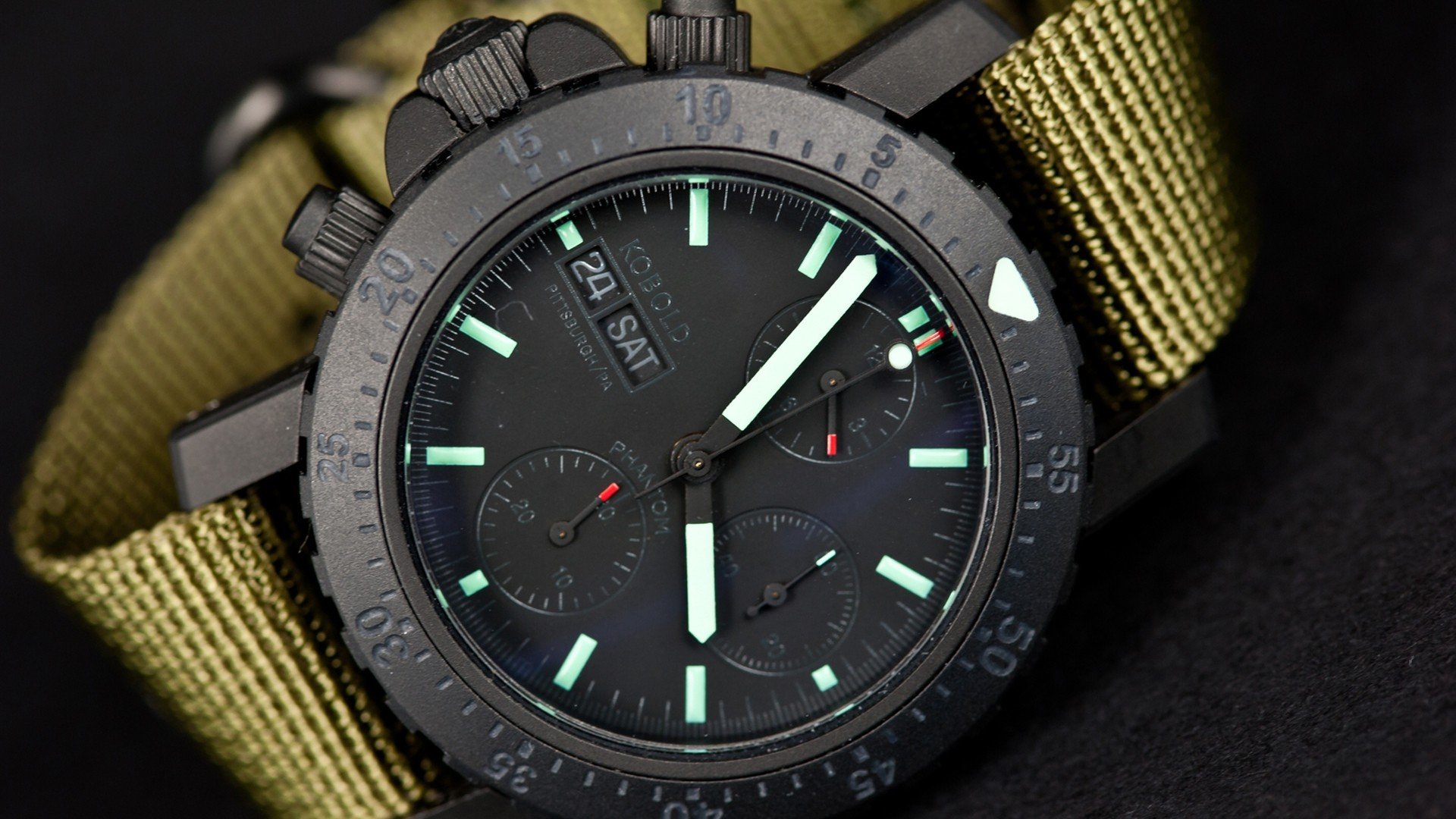 Close-up HD desktop wallpaper of a man-made black chronograph watch with glowing hands and a textured olive green fabric strap.