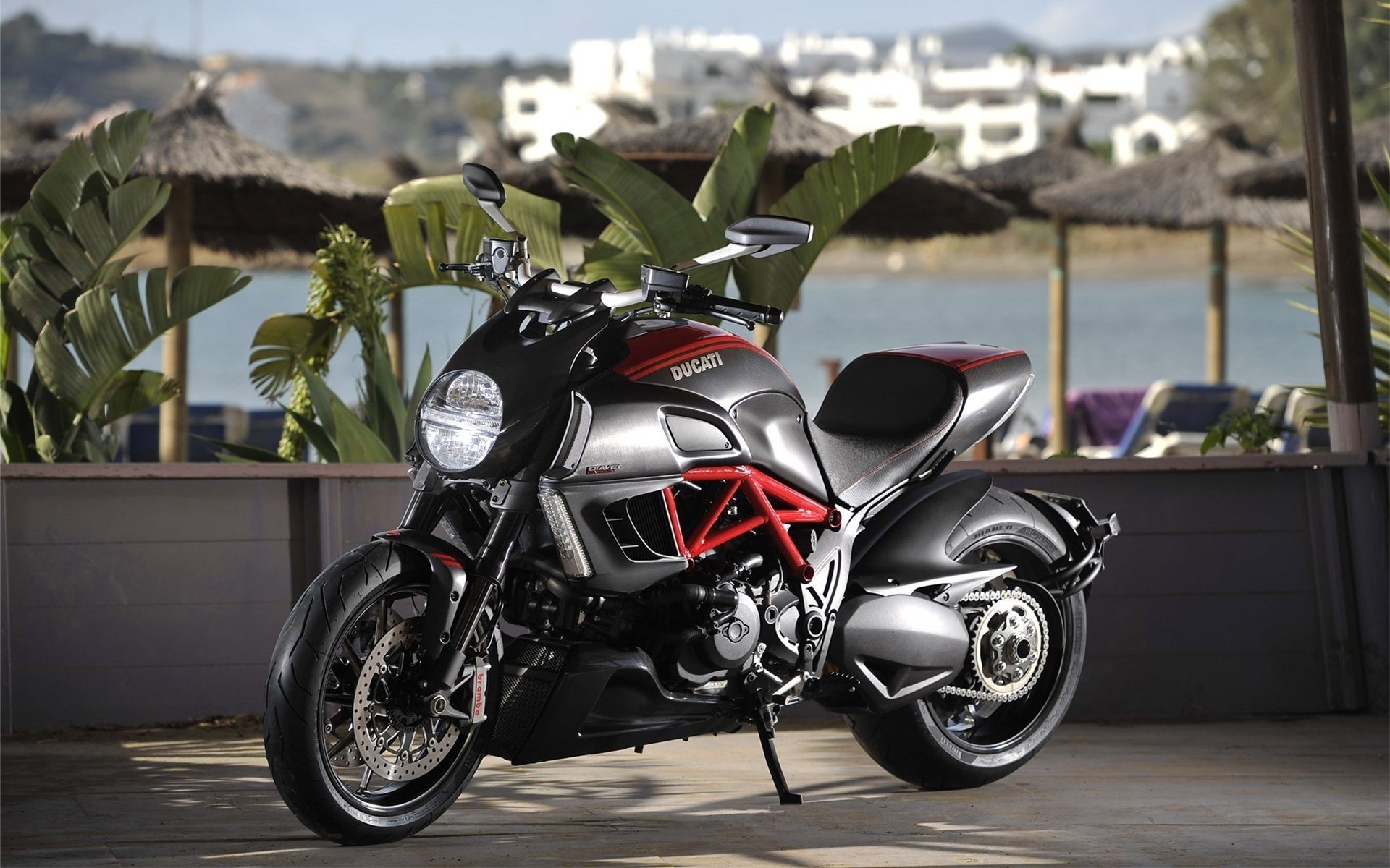 HD PC desktop wallpaper showcasing a sleek Ducati motorcycle parked indoors with a scenic waterfront and white buildings in the blurred background.