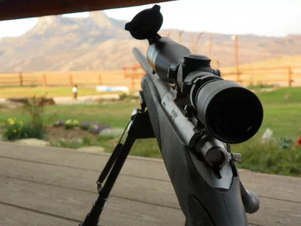 4K Ultra HD PC desktop wallpaper: man-made sniper rifle on a bipod, close-up of scope and barrel aimed over a sunlit rural landscape.