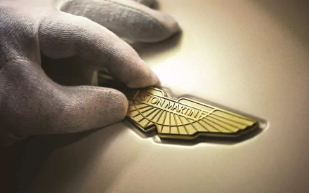 Close-up of a gloved hand holding an Aston Martin emblem, captured in high definition for a sleek PC desktop wallpaper background.