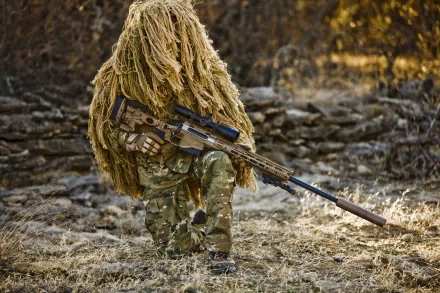 A camouflaged sniper crouches in a natural setting, holding a high-precision rifle, embodying military expertise. This HD wallpaper captures the stealth and skill of modern warfare.