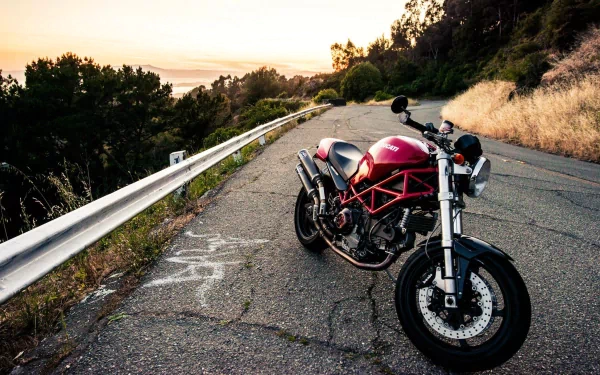 HD PC desktop wallpaper featuring a red Ducati motorcycle parked on a scenic winding road at sunset with trees and guardrails in the background.