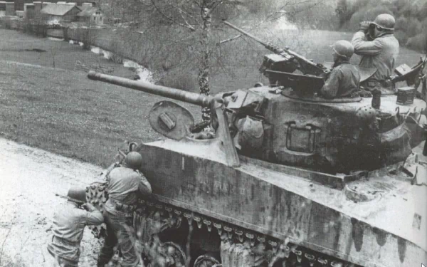 Black-and-white HD PC desktop wallpaper of a military M4 Sherman tank with soldiers aboard and beside it, rolling along a rural road past village houses.