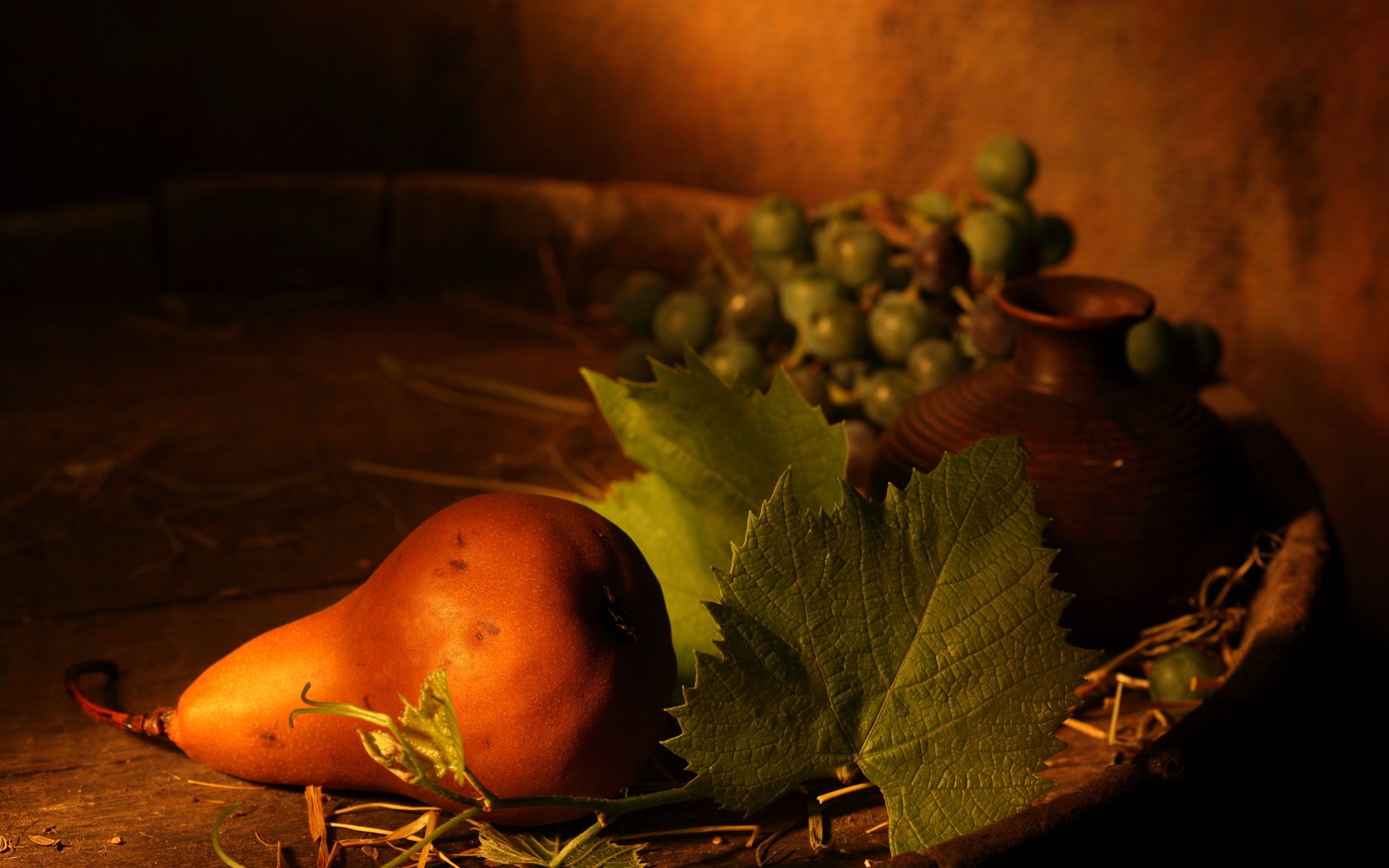 HD still life desktop wallpaper featuring a ripe pear, grape leaves, green grapes, and a clay pot arranged on a rustic wooden surface with warm lighting.
