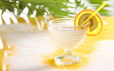 HD desktop wallpaper featuring a refreshing cocktail in a clear glass with a yellow straw and citrus garnish, set against a sunlit tropical background with green leaves.