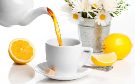 A teapot pours tea into a white cup, accompanied by slices of lemon and a sweetener on a plate. A vase of daisies adds a fresh touch to this inviting food and tea scene.