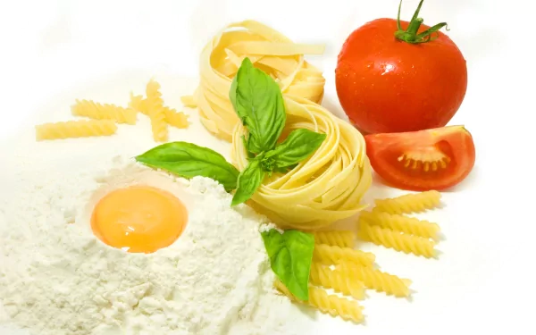 A vibrant arrangement of fresh ingredients for pasta, featuring fettuccine, flour, an egg, basil, and ripe tomatoes, creating an appetizing food-themed HD desktop wallpaper.